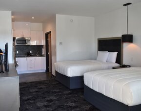 A guest room with two beds dressed in white bedding, black headboards, bedside tables, pendant lighting and a kitchenette in the background at Hotel Bella.