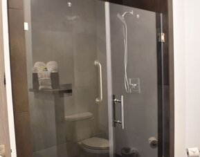 A modern bathroom with a glass-enclosed walk-in shower, dark grey wall tiles, chrome fixtures and a built-in shelf holding folded towels at Hotel Bella.