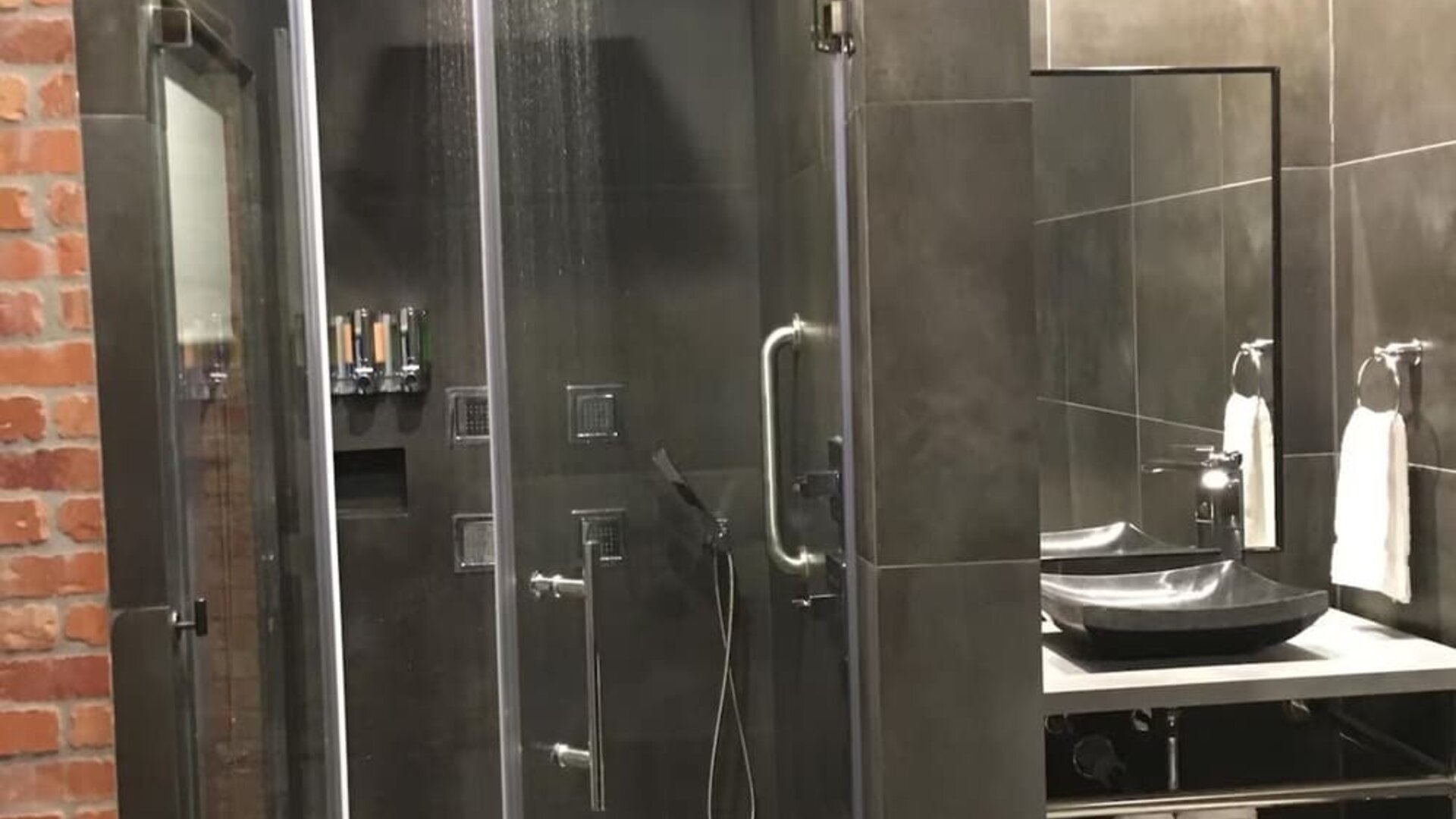 A walk-in shower with glass doors, dark grey tiles, a rainfall showerhead, handheld shower fixture and a wall-mounted shelf at Hotel Bella.
