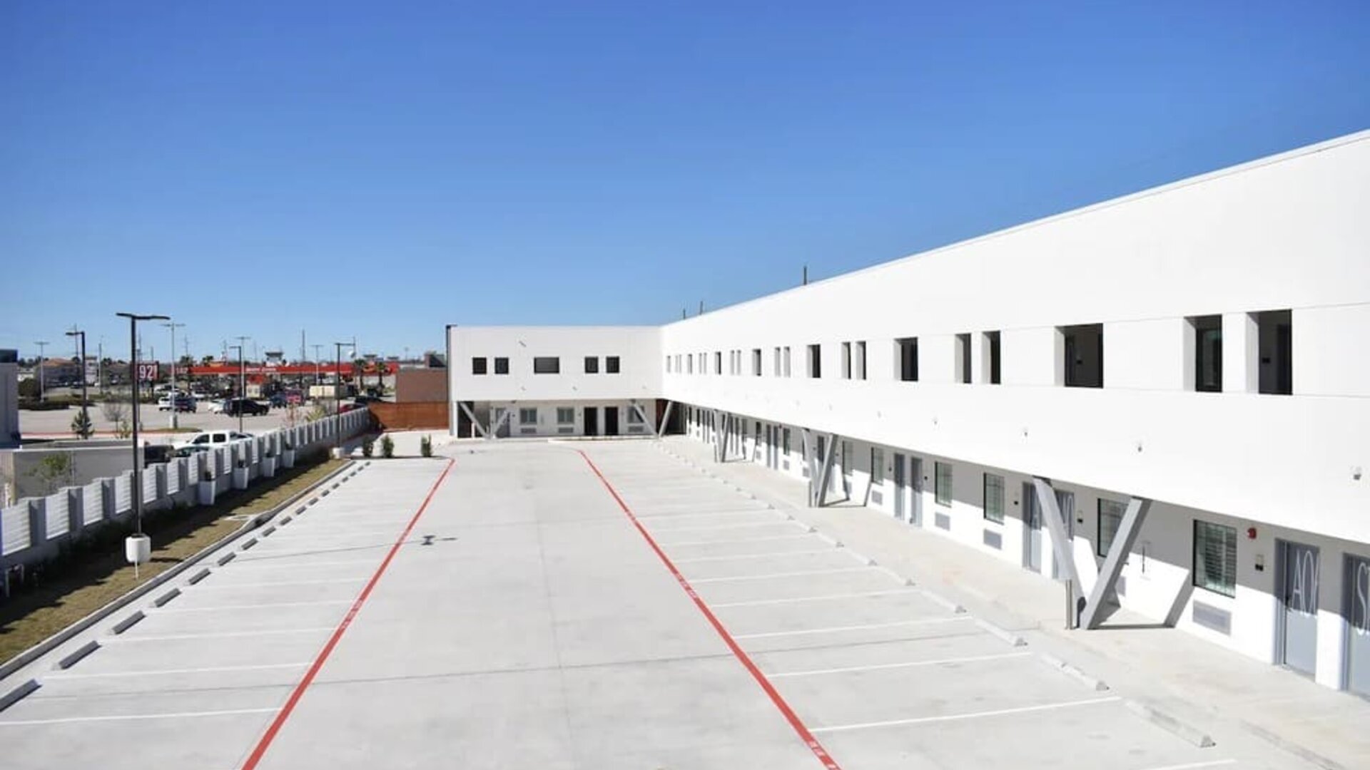 A wide exterior view of the modern white hotel building, showing the open-air corridors, numbered rooms and large concrete parking area at Hotel Bella.