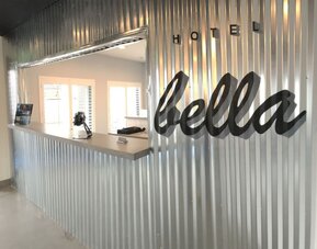 A hotel reception desk set against a corrugated metal wall, featuring a large script-style hotel sign and a bright, minimalist reception area at Hotel Bella.