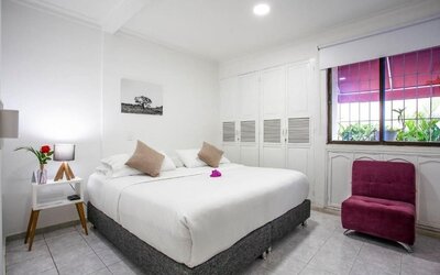 A bright guest room featuring a king-size bed with white linens, beige accent cushions, a magenta upholstered armchair, built-in white wardrobes and a large window overlooking greenery at Mucura Hotel And Spa.