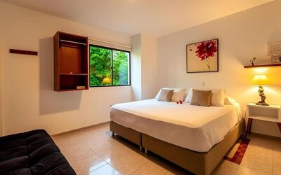 A spacious guest room featuring a king-size bed with white linens and beige cushions, a black sofa, a wall-mounted television, wooden shelving and a large window overlooking greenery at Mucura Hotel And Spa.