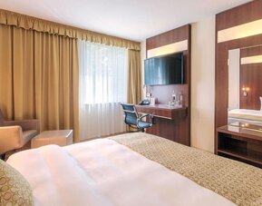 A guest bedroom with a king bed dressed in white linens and patterned bedding, warm wood-panelled furniture, a desk and chair, a large wall-mounted television and a comfortable seating area at Best Western Macrander Hotel Frankfurt/Kaiserlei.