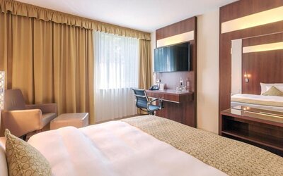 A guest bedroom with a king bed dressed in white linens and patterned bedding, warm wood-panelled furniture, a desk and chair, a large wall-mounted television and a comfortable seating area at Best Western Macrander Hotel Frankfurt/Kaiserlei.