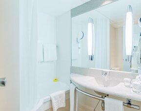 A bright, modern hotel bathroom with a bathtub and shower, a wide vanity basin, a large mirror, wall-mounted lighting, fresh towels and complimentary toiletries at Best Western Macrander Hotel Frankfurt/Kaiserlei.