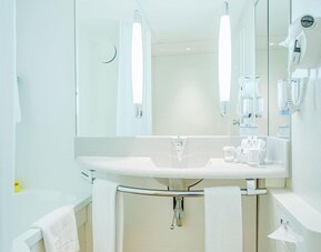 A spacious bathroom with a white bathtub, a curved vanity basin, an expansive mirror, contemporary lighting, a wall-mounted hairdryer and white towels, complemented by warm wood-effect flooring at Best Western Macrander Hotel Frankfurt/Kaiserlei.