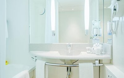A spacious bathroom with a white bathtub, a curved vanity basin, an expansive mirror, contemporary lighting, a wall-mounted hairdryer and white towels, complemented by warm wood-effect flooring at Best Western Macrander Hotel Frankfurt/Kaiserlei.