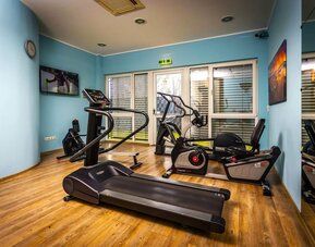 The hotel's fitness room, equipped with treadmills, stationary exercise bikes and other cardio equipment, with mirrored walls, blue accents and wood-effect flooring at Best Western Macrander Hotel Frankfurt/Kaiserlei.