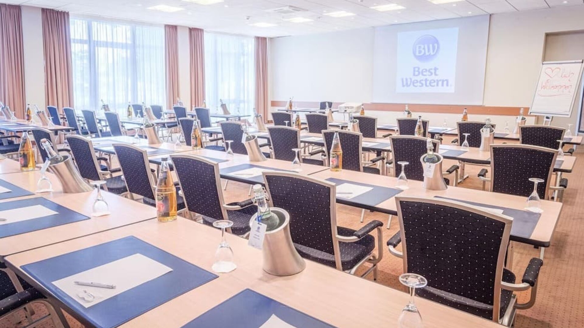 A conference room arranged for meetings and presentations, with rows of tables and chairs, writing materials, water bottles, presentation equipment and a projection screen displaying the Best Western logo.