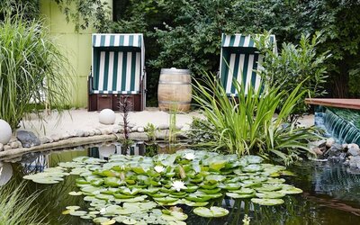 A peaceful landscaped garden with a small pond covered in lily pads and water lilies, surrounded by lush greenery and two striped outdoor deckchairs.