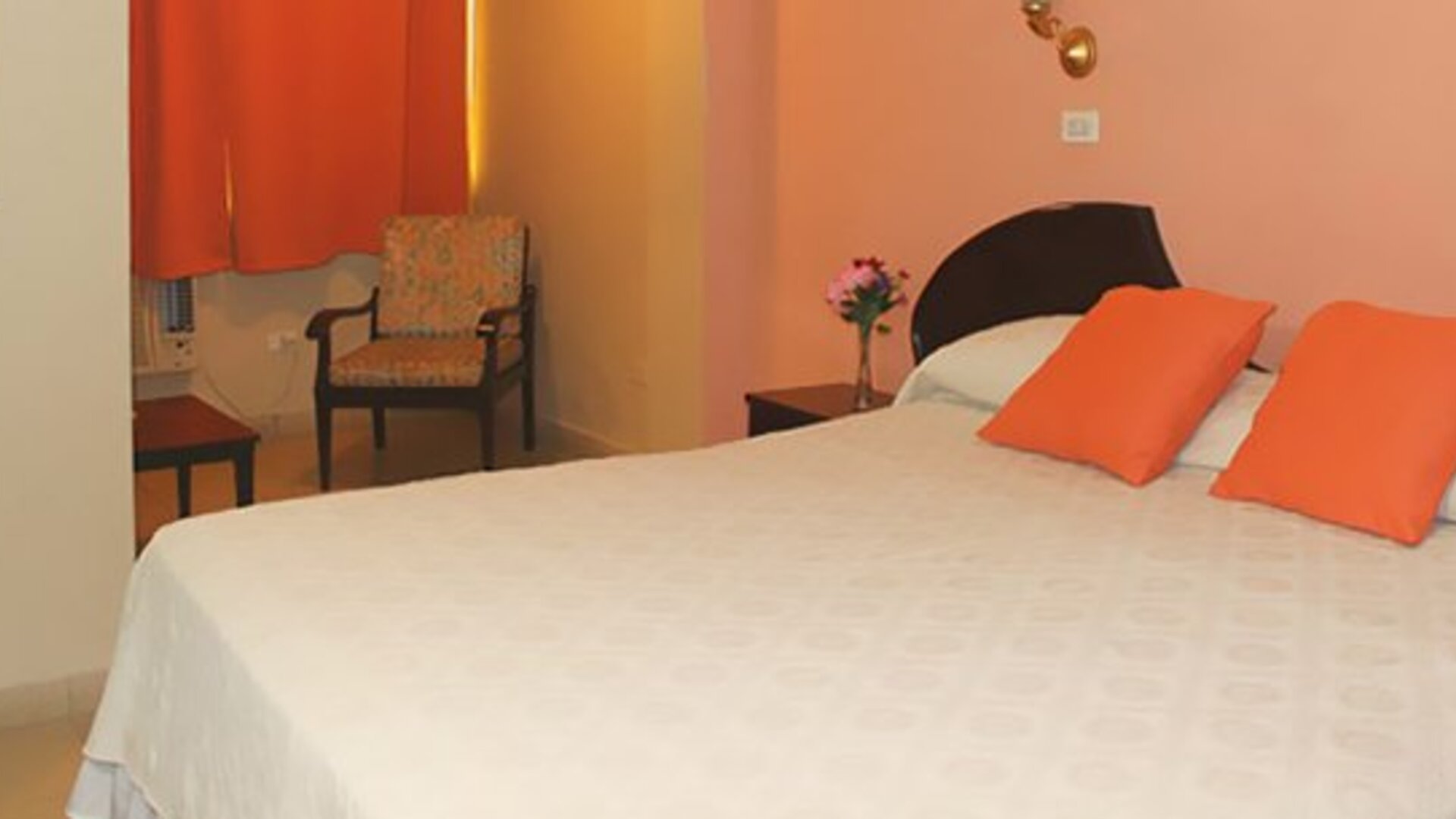 White bed with orange cushions, peach walls, orange curtains and a patterned armchair at Caribe Panamá Hotel.