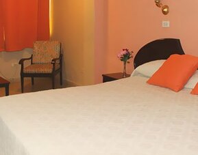 White bed with orange cushions, peach walls, orange curtains and a patterned armchair at Caribe Panamá Hotel.
