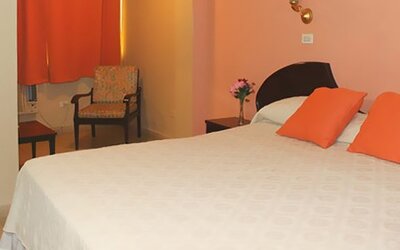 White bed with orange cushions, peach walls, orange curtains and a patterned armchair at Caribe Panamá Hotel.