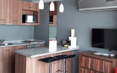 Modern kitchenette and living area with wood cabinetry, microwave, granite breakfast bar, bar stools and wall-mounted television at Hotel Block Suites.