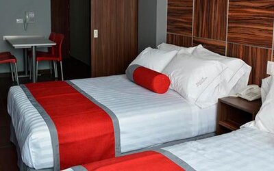 Twin beds with white striped bedding, red runners and cylindrical cushions, dark wood headboard and a small dining table at Hotel Block Suites.