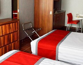 Guest room with two beds featuring white striped bedding, red runners, wood-finish furniture, wall-mounted television and a small dining area at Hotel Block Suites.