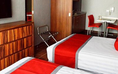 Guest room with two beds featuring white striped bedding, red runners, wood-finish furniture, wall-mounted television and a small dining area at Hotel Block Suites.