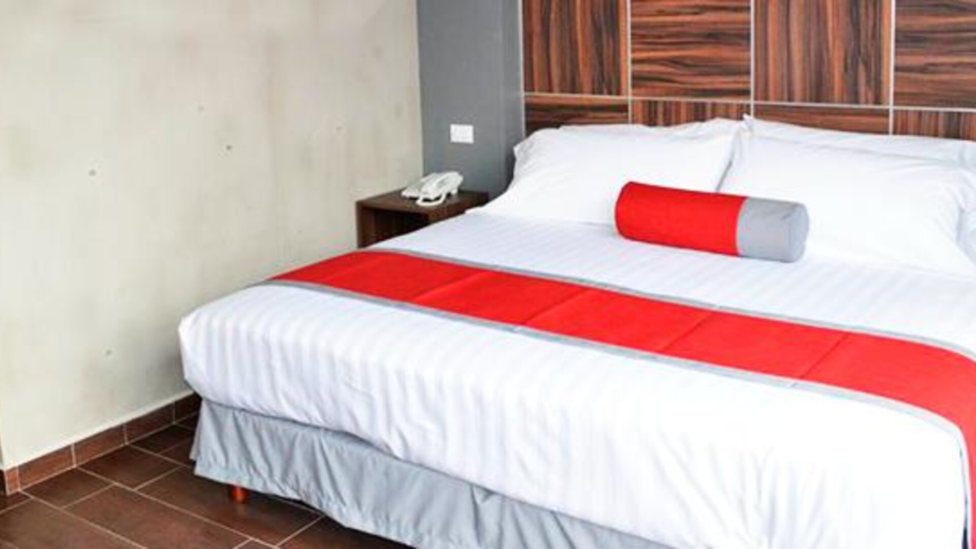 King-size bed with white striped bedding, red bed runner and cylindrical accent cushion, dark wood headboard and bedside tables at Hotel Block Suites.