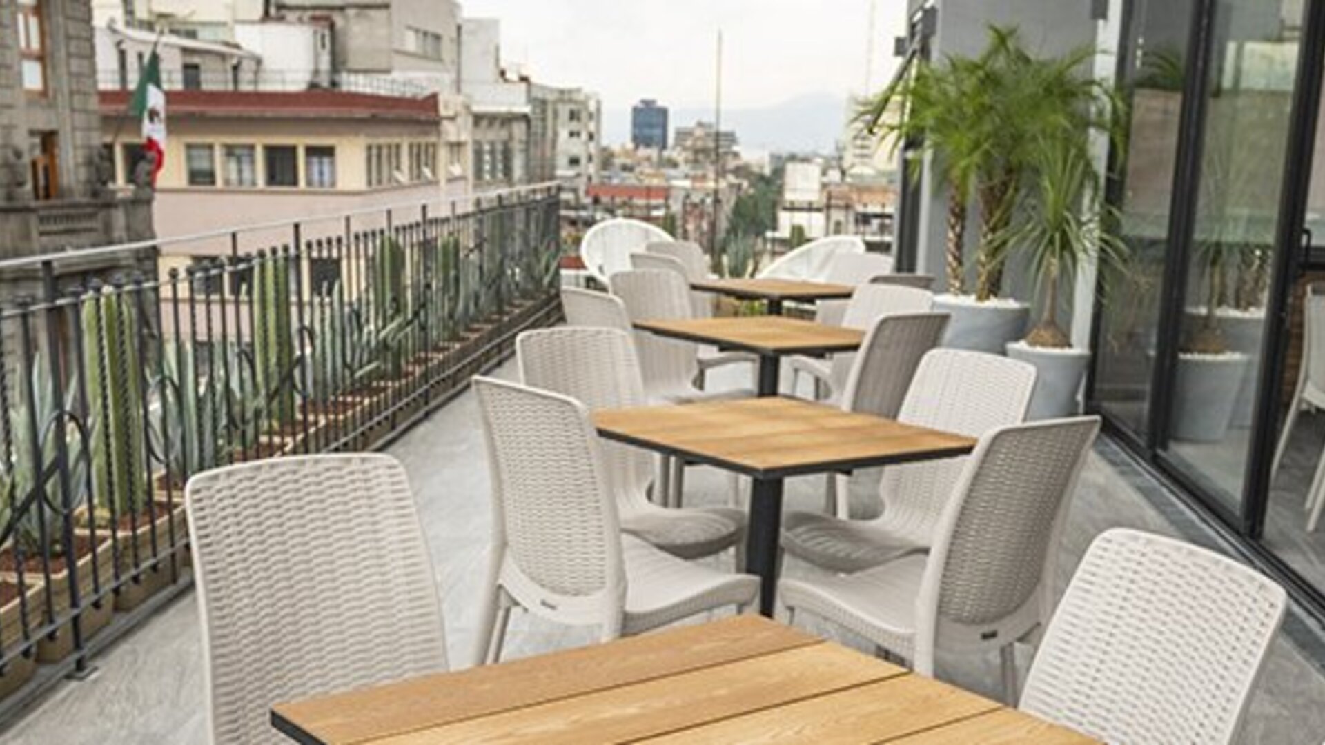 Outdoor terrace dining area with wooden tables, white woven chairs and city views at Hotel Principal.