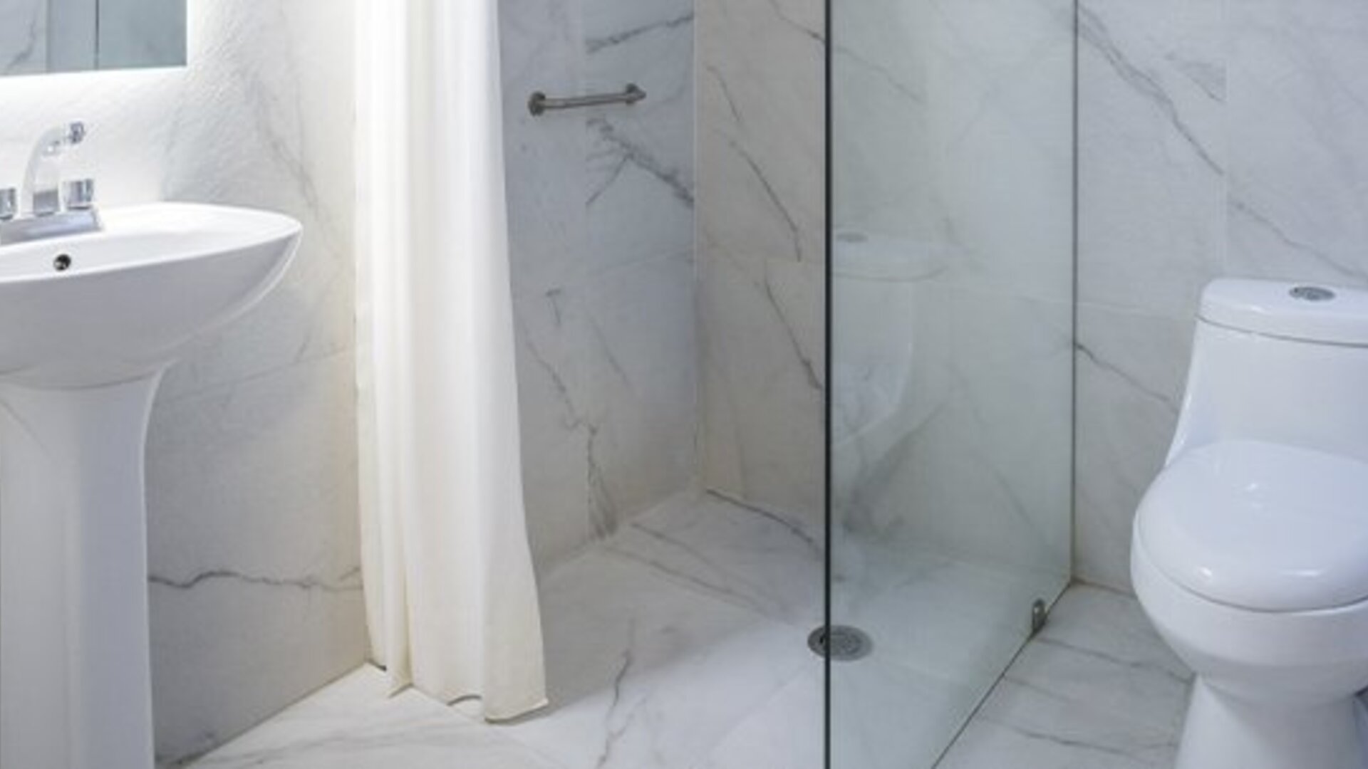 Bathroom with white marble-effect tiles, pedestal sink, glass-enclosed shower and toilet at Hotel Principal.
