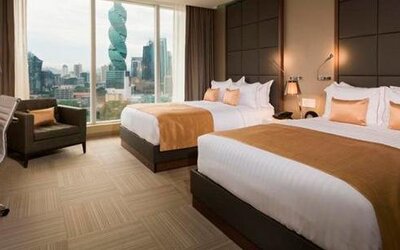 Guest room with two double beds, gold accent cushions, an armchair and city views through large windows at Hotel Las Américas Golden Tower Panamá.