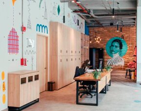 Shared workspace with a long wooden table, chairs, lockers, colourful wall illustrations and exposed concrete columns at UMA Suites Chía.