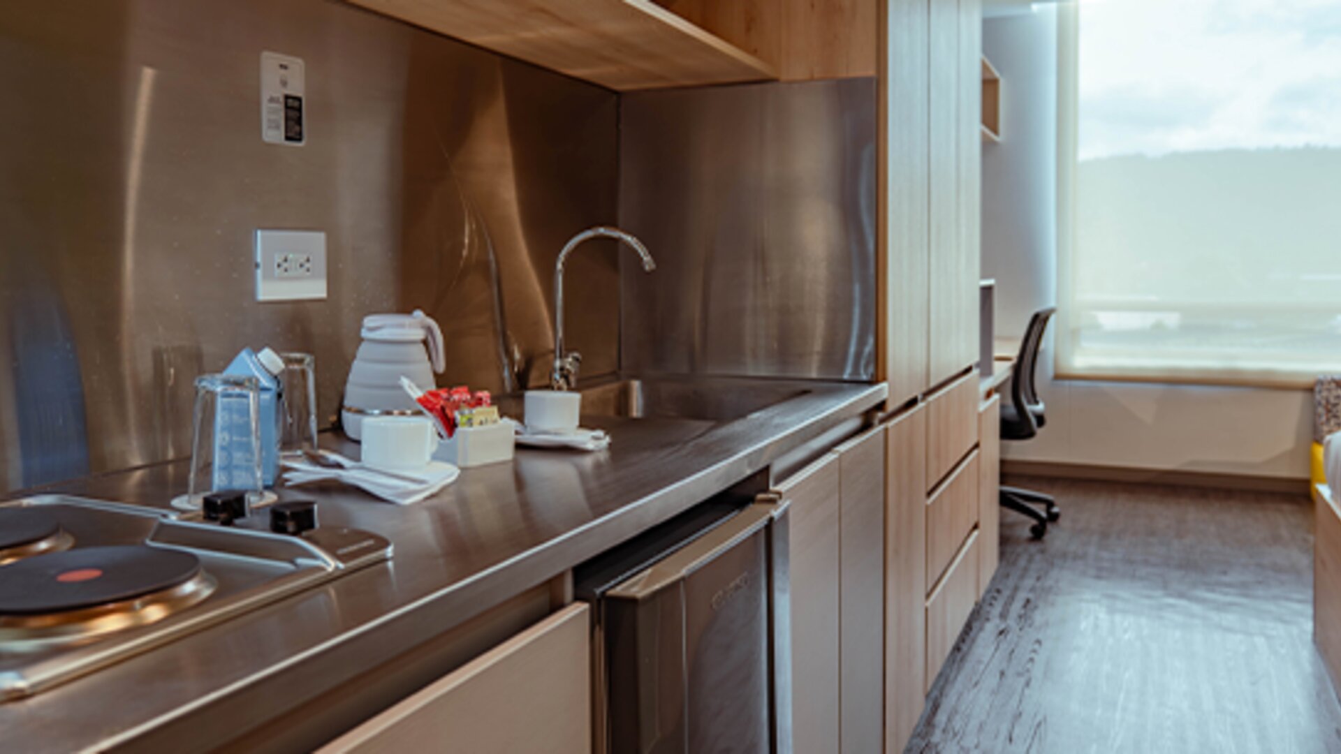 Studio kitchenette with a two-burner electric hob, stainless-steel backsplash, sink, kettle and wooden cabinetry at UMA Suites Chía.