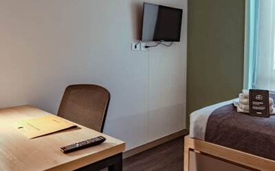 Compact guest room with a single bed, dark bed runner, wooden desk, chair and wall-mounted television at UMA Suites Chía.
