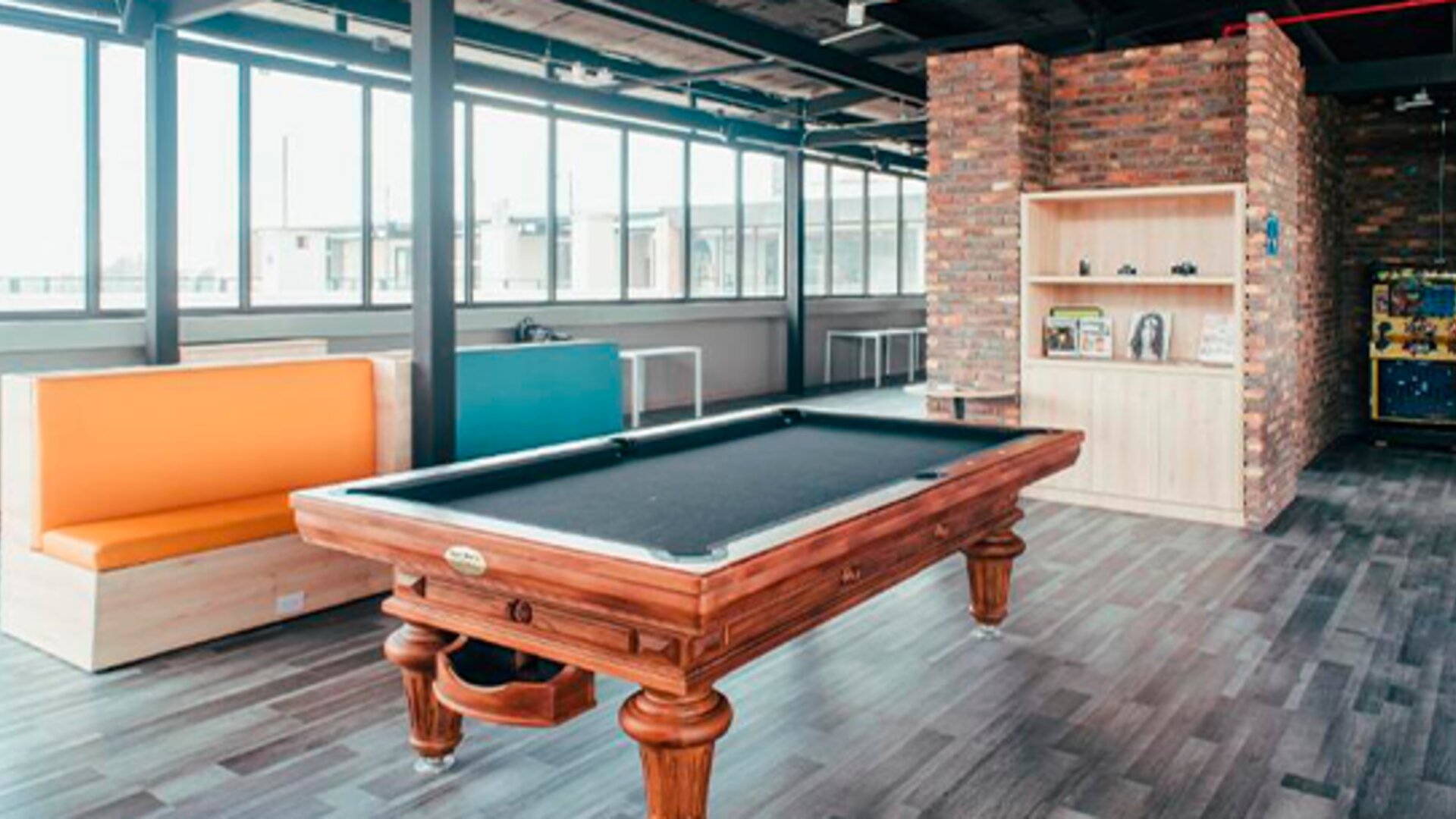 Games room with a wooden pool table, colourful booth seating, large windows and exposed brickwork at UMA Suites Chía.