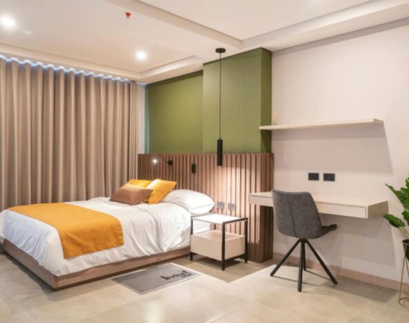 Guest bedroom with a double bed, mustard and brown cushions, green accent wall, wooden slat headboard, desk, and potted plant at Hotel Living 35 Suites.