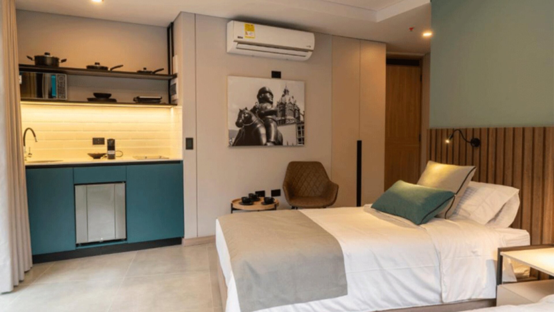 Guest suite with a double bed, teal kitchenette cabinetry, wooden headboard, wall-mounted television, and compact seating area at Hotel Living 35 Suites.