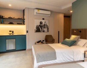 Guest suite with a double bed, teal kitchenette cabinetry, wooden headboard, wall-mounted television, and compact seating area at Hotel Living 35 Suites.