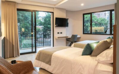 Guest bedroom with two beds, neutral bedding, teal and green accent cushions, a wall-mounted television, desk, and balcony doors at Hotel Living 35 Suites.