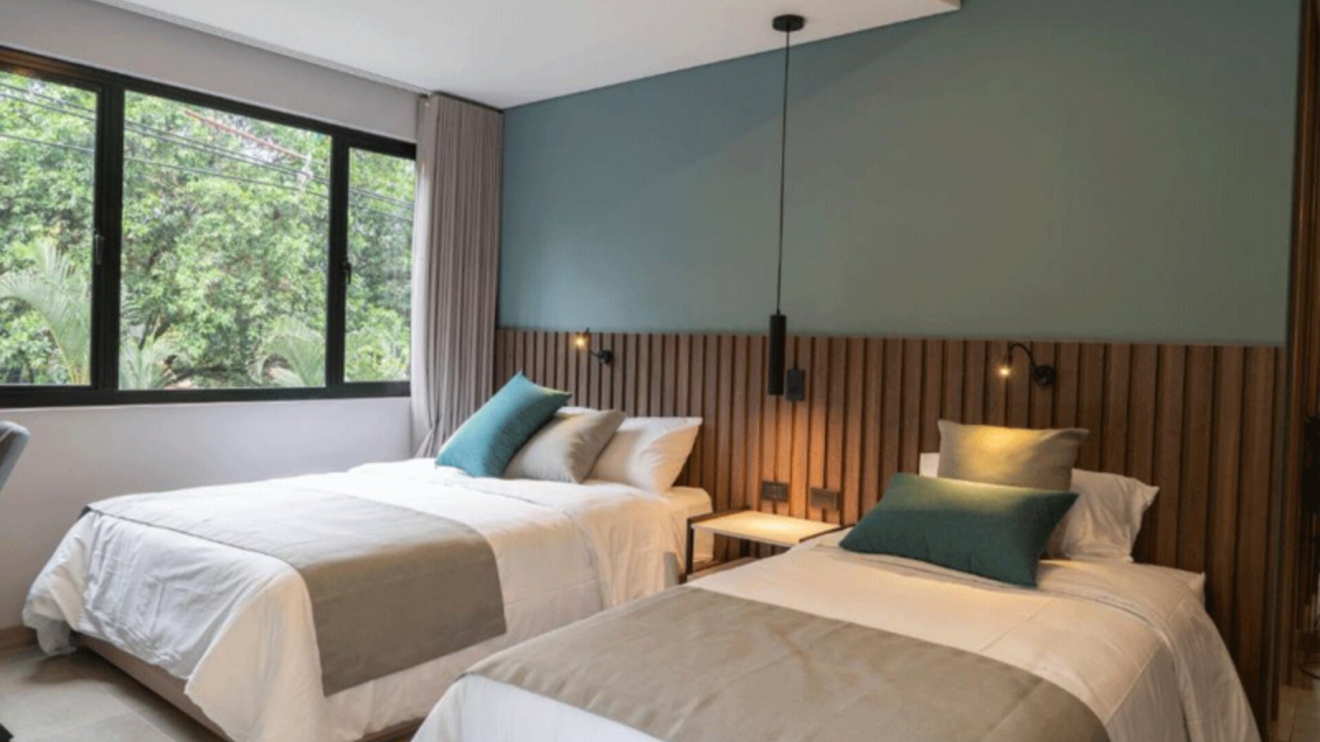 Guest room with two beds dressed in white linens, teal accent cushions, a wooden slat headboard, and large windows at Hotel Living 35 Suites.