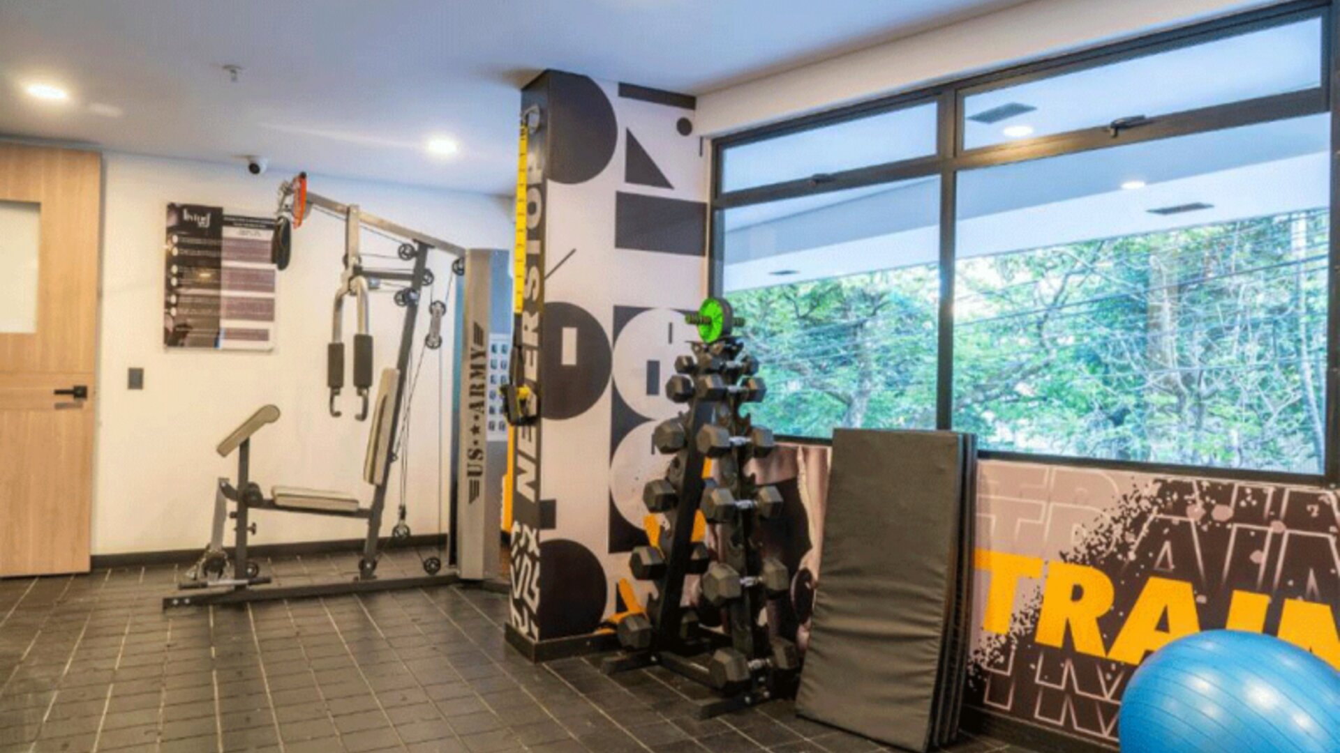 Fitness room with strength-training equipment, dumbbells, exercise mats, a stability ball, and large windows at Hotel Living 35 Suites.