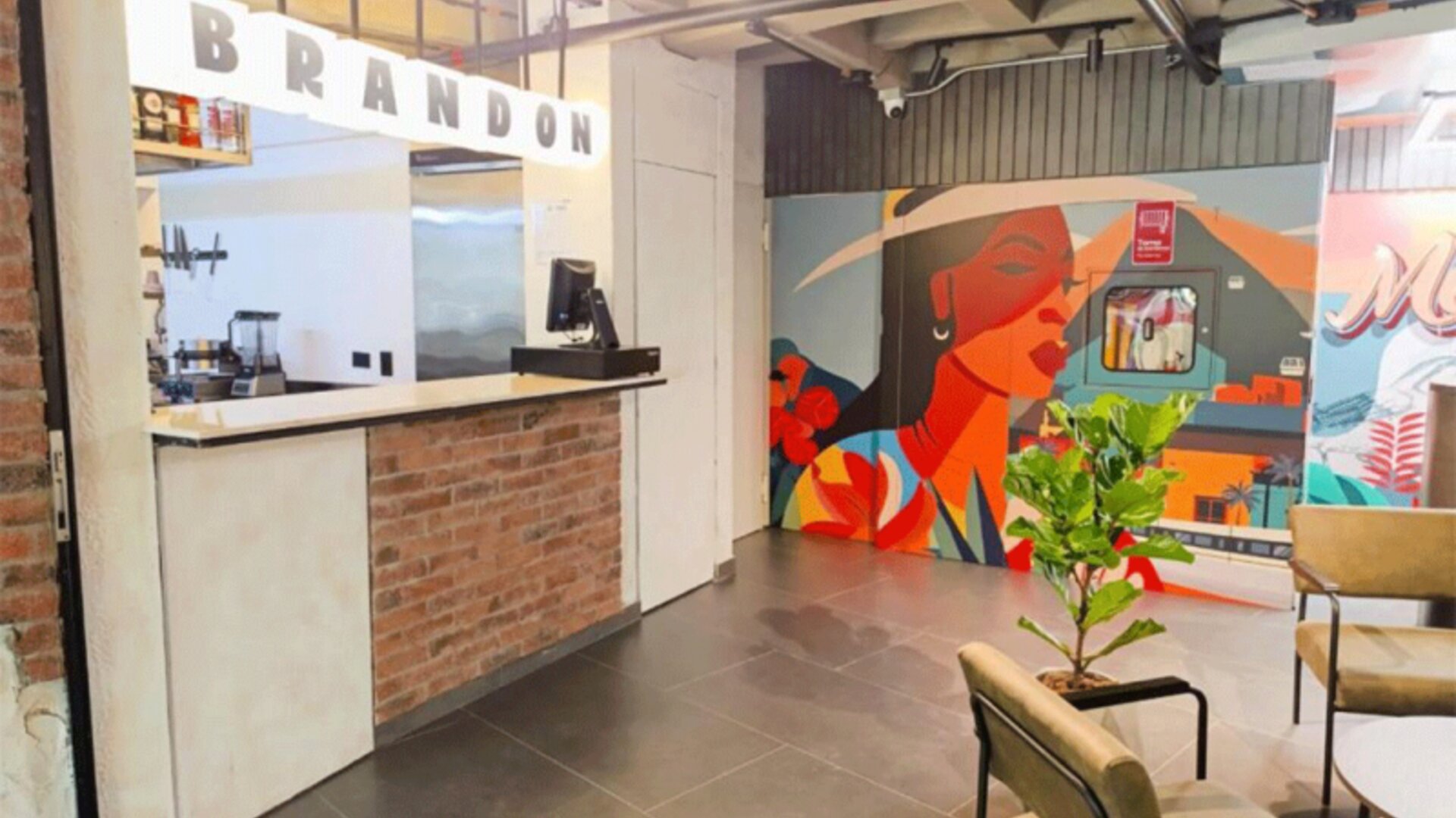 Reception counter with a brick-front desk, illuminated BRANDON lettering, colourful mural artwork, and indoor plants at Hotel Living 35 Suites.