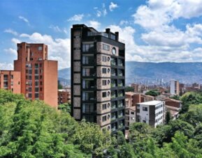 Exterior view of a multi-storey dark-brick hotel building surrounded by green trees, with city buildings and mountains in the background at Hotel Living 35 Suites.