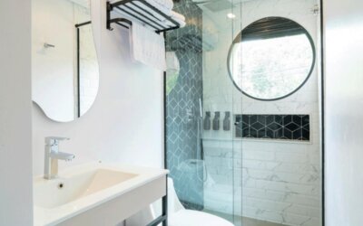 Modern bathroom with a white vanity, curved mirror, glass-enclosed shower, white and grey patterned tiles, and a round window at Hotel Living 35 Suites.