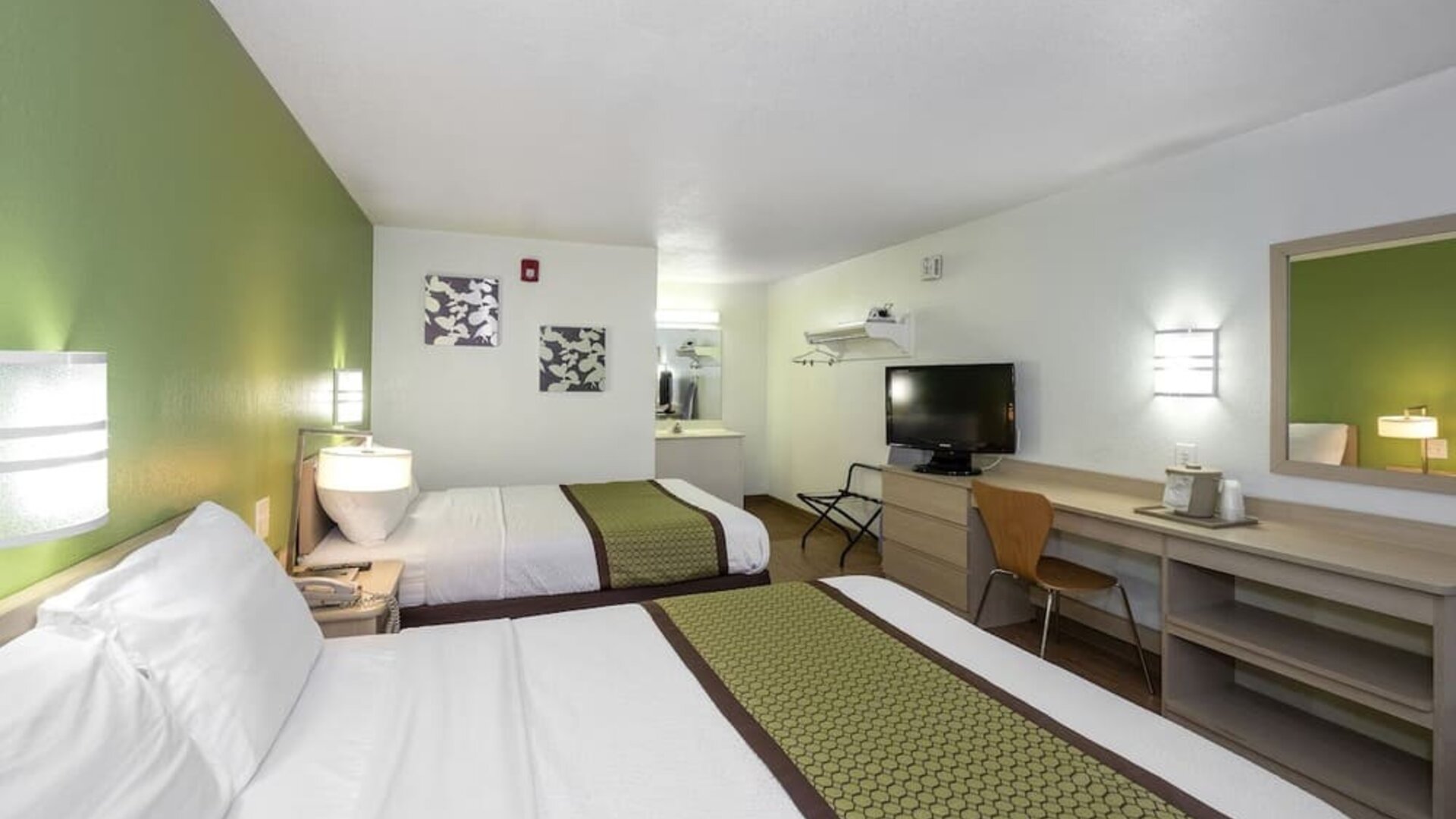 Guest room with two double beds, white bedding, green patterned runners, bedside lighting, a television, and a long desk with a mirror at American Inn Carlisle.