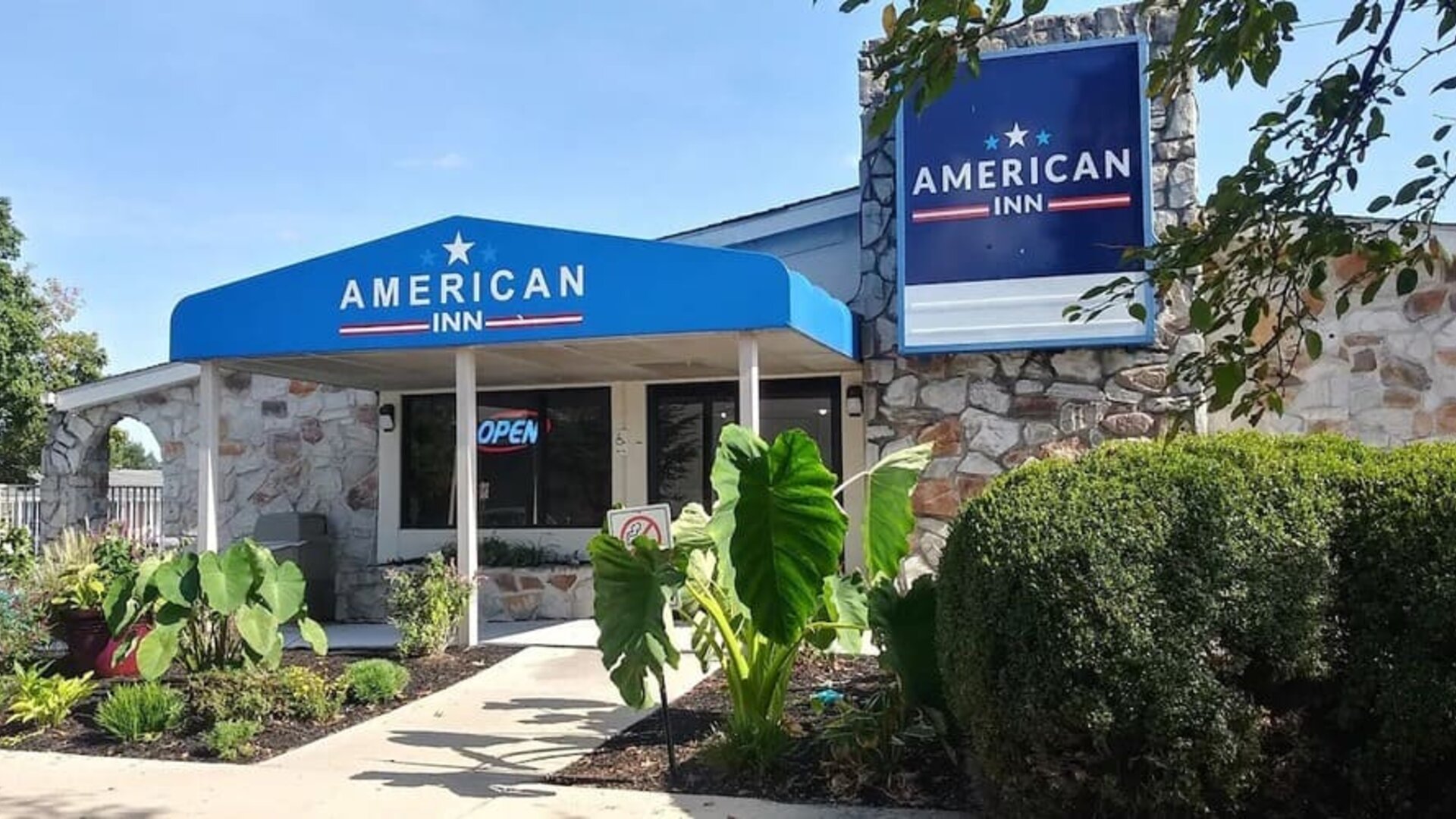Exterior entrance of American Inn Carlisle featuring a blue entrance canopy, hotel signage, stone façade, landscaped plants, and shrubs.