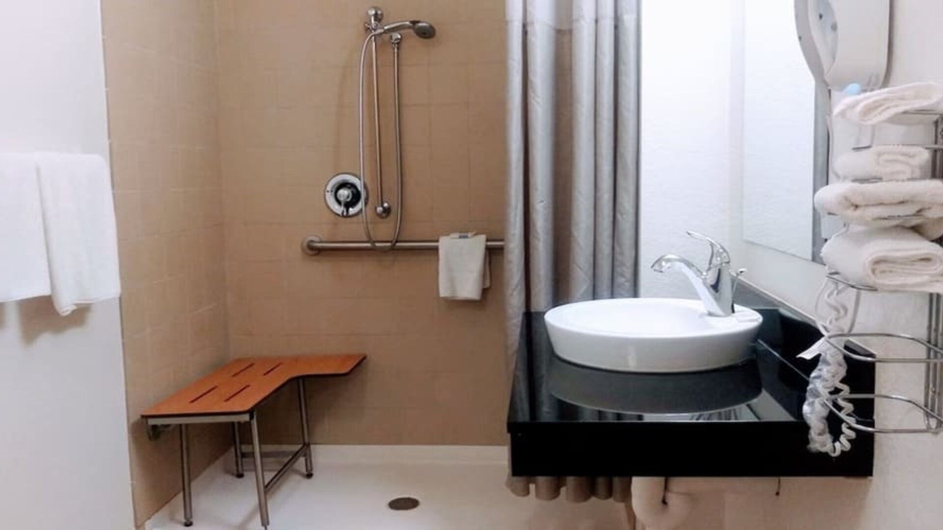 Bathroom with a walk-in shower, grab bars, shower seat, white vessel sink, black countertop, and folded towels at American Inn Carlisle.