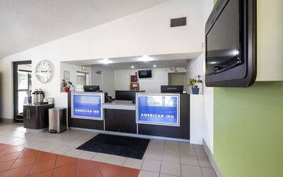Reception desk with American Inn Carlisle signage, computers, a wall-mounted television, and a tiled lobby floor.