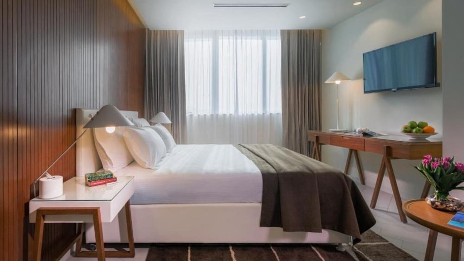 Bright guest bedroom with a king-size bed, white bedding, dark throw, wood-panelled wall, bedside table, television, and desk at Prima Millennium.