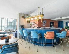 Modern hotel bar with a curved blue counter, colourful upholstered bar stools, pendant lighting, and lounge seating at Prima Millennium.