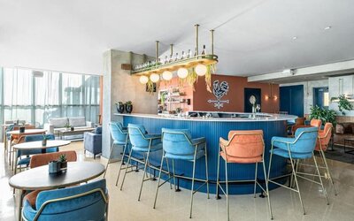 Modern hotel bar with a curved blue counter, colourful upholstered bar stools, pendant lighting, and lounge seating at Prima Millennium.