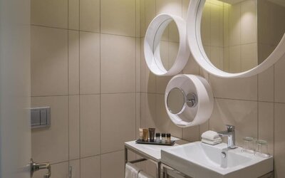 Contemporary bathroom with beige tiled walls, a white vanity sink, circular mirrors, toiletries, and neatly folded towels at Prima Millennium.