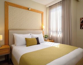 Guest room featuring a king-size bed with white bedding, a gold bed runner, decorative accent cushions, wooden bedside tables, and a large upholstered headboard at Prima Park Hotel.