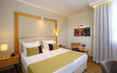 Guest room with a king-size bed, white bedding, gold and dark accent cushions, a wooden headboard frame, and a small seating area beside the window at Prima Park Hotel.