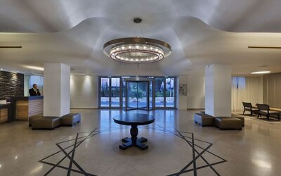 Hotel reception lobby with a front desk, polished flooring, a circular chandelier, seating areas, and glass entrance doors at Prima Park Hotel.